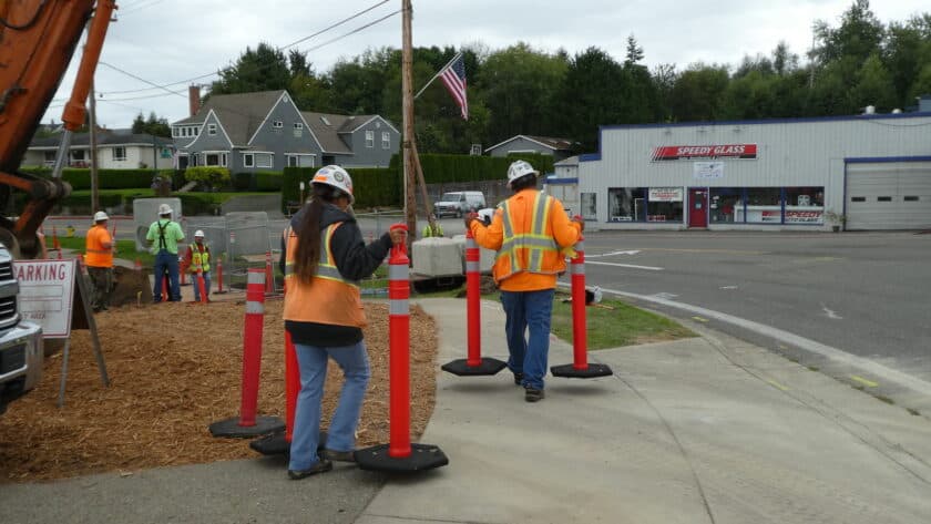 Workers carry orange traffic barriers on the Harborview Drive sidewalk where utilities are being moved for a new roundabout.