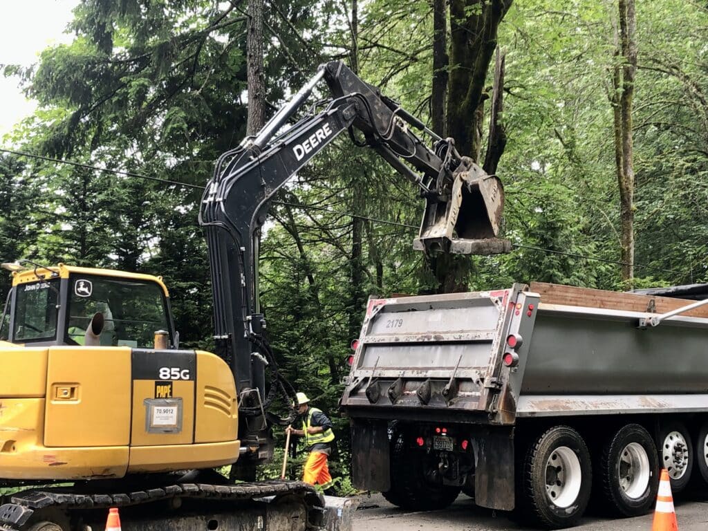 An escavator loads a cut-up piece of concrete roadway into a dump truck.