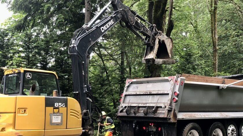 An escavator loads a cut-up piece of concrete roadway into a dump truck.