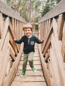 Boy running over a wobbly wooden chain bridge at park.