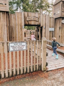 A boy and a girl run into a wooden fort entrance with tall planks and beams leading to a playground