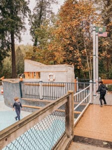 Two children run around a boat structure at a playground under tall trees