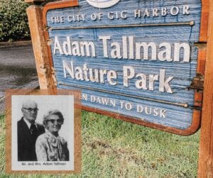 This is a photo of a wooden sign painted blue with the words "Adam Tallman Nature Park" on it. The photo has a newspaper clipping overlay of an older couple in the corner featuring Adam Tallman.