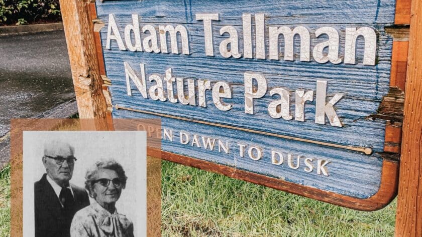 This is a photo of a wooden sign painted blue with the words "Adam Tallman Nature Park" on it. The photo has a newspaper clipping overlay of an older couple in the corner featuring Adam Tallman.