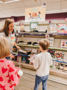 woman hands out paper booklets to two kids next to a bookshelf