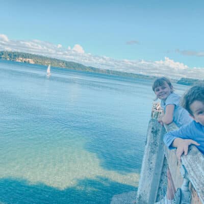This is a photo of two kids posing behind a wooden railing above the water. A sailboat is in the background.