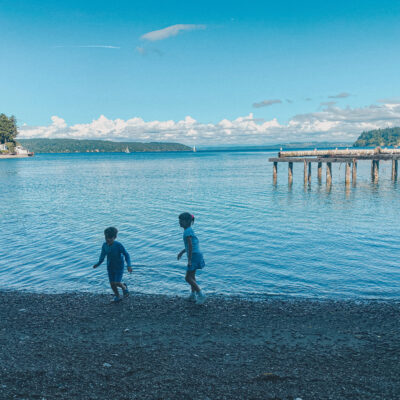 This is a photo of the silhouette of two children playing at the beach. The water is blue and there's a bluff covered in trees in the background.