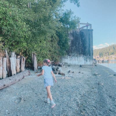 This is a photo of a little girl walking on a sandy beach. There's a green tree or bush and a concrete landing behind her. The water is blue and there's a bluff covered in trees in the background.