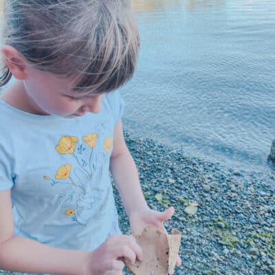 This is a photo of a little girl looking at a brown piece of paper she found on a sandy beach.