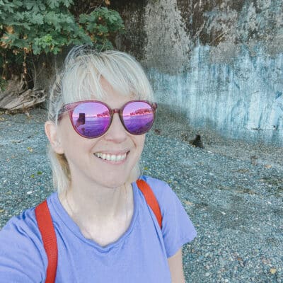 This is a photo of a mom with purple sunglasses and a purple t-shirt standing on a sandy beach taking a selfie. She's super cool.